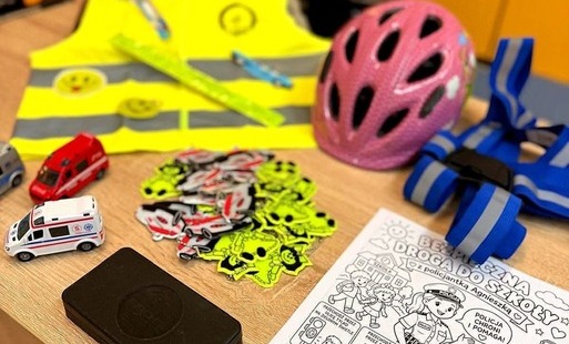 **Tekst alternatywny:**\  Materiały edukacyjne dotyczące bezpieczeństwa dzieci na drodze, w tym odblaski, kamizelki, kask rowerowy, samochodziki oraz karta pracy „Bezpieczna droga do szkoły”